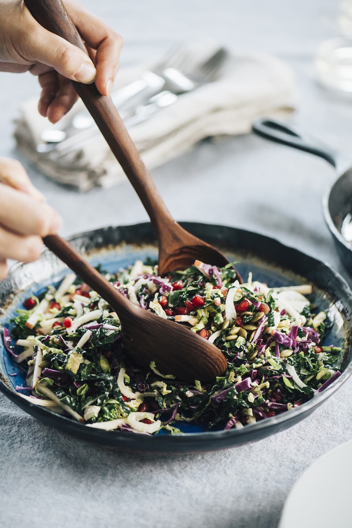 A woman's hands tossing kale pomegranate salad in a blue ceramic bowl.