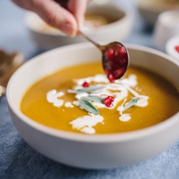 A bowl of roasted butternut squash soup being garnished with pomegranate seeds.