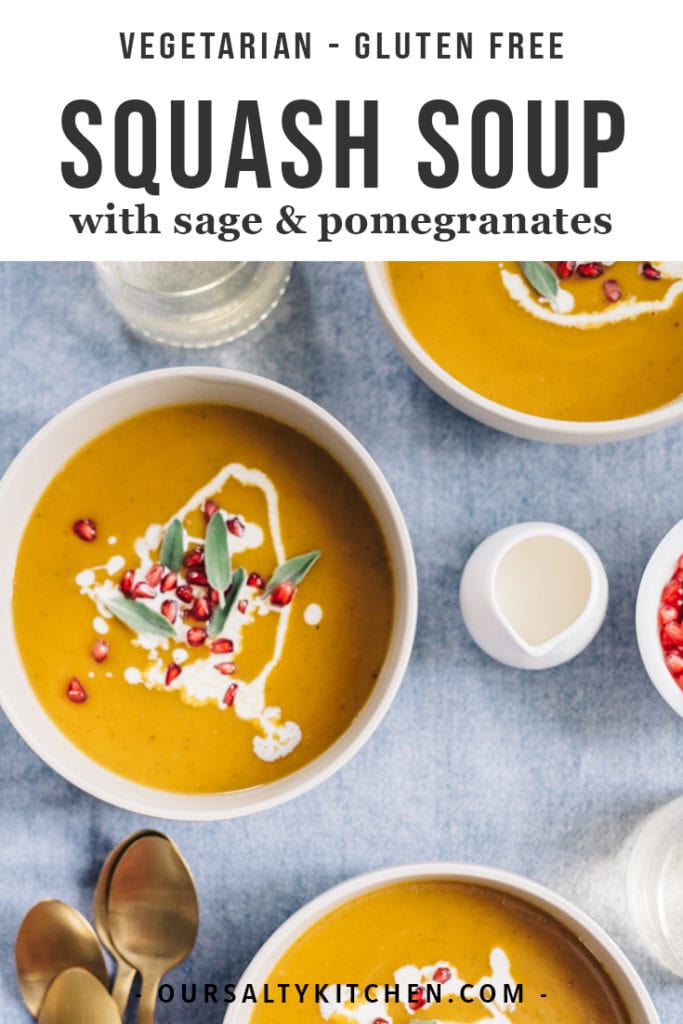 Two bowls of butternut squash soup garnished with pomegranate seeds, sage, and fresh cream on a blue tablecloth.