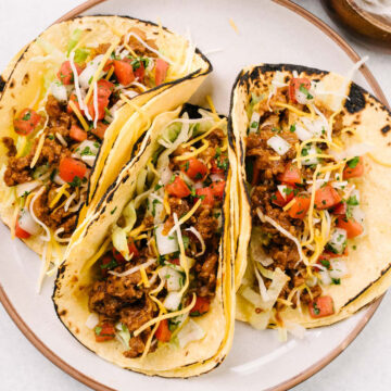 Three ground beef tacos on a cream colored plate topped with pico de gallo, shredded cheese, and shredded lettuce; small bowls of pico de gallo and sour cream to the side.