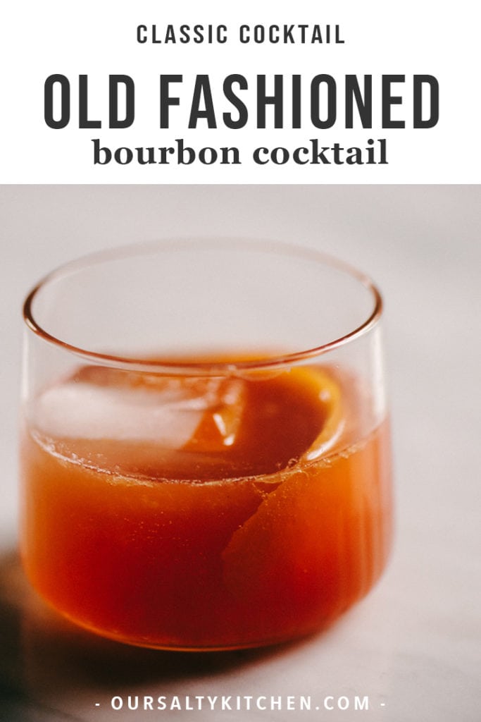 A classic old fashioned bourbon cocktail in a cocktail glass with a giant ice cube and orange peel garnish.