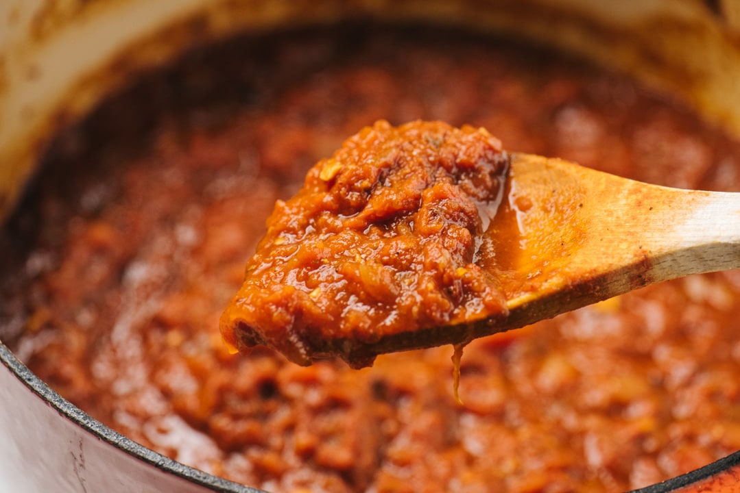 A wood spoon covered in slow simmered sunday sauce hovering over a pot on the stove.