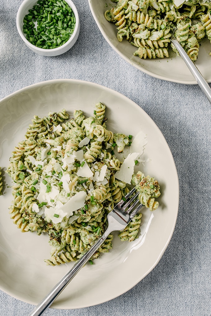 A plate of whole wheat pasta tossed with roasted cauliflower and creamy ricotta spinach pesto, garnished with fresh chives and shaved parmesan cheese. 