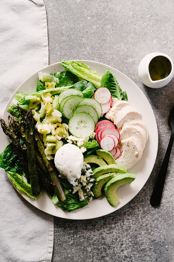 Spring cobb salad with chicken, asparagus, leeks, avocado, and blue cheese over romaine lettuce on a cream plate with a side of vinaigrette in a small pitcher.
