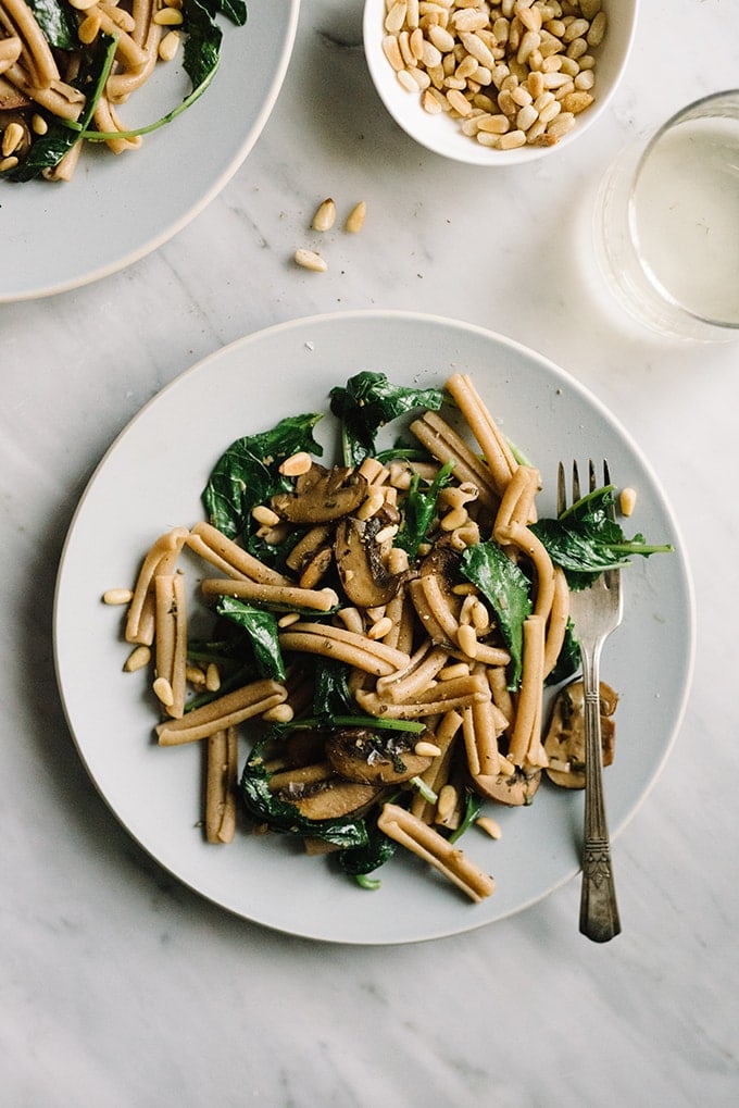 A serving of kale pasta on a blue plate  alongside a glass of wine wine and a small dish of toasted pine nuts. 