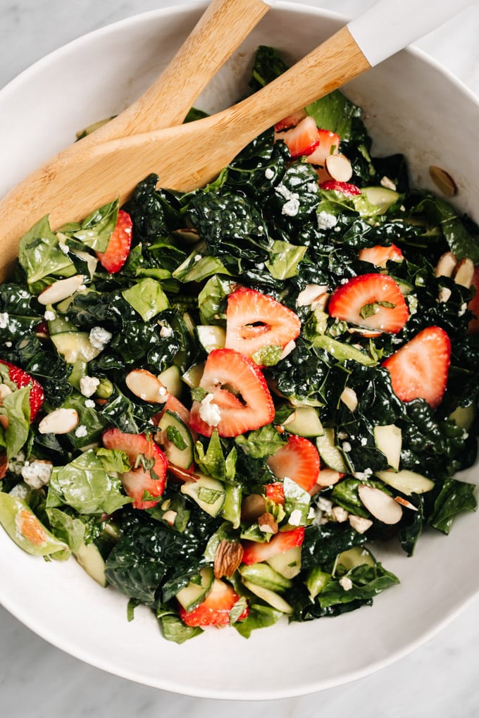 A large white serving bowl filled with strawberry kale salad and wood serving spoons.