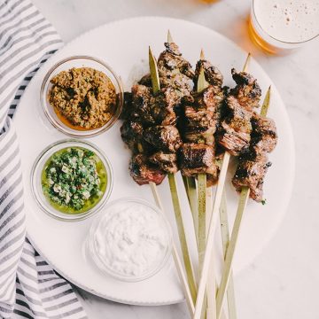 These paleo grilled flank steak skewers are a great party food recipe! Thin strips of grass fed flank steak is charred and crispy on the outside, and and tender on the inside. A real food dipping sauce trio of chimichurri, minty yogurt, and sundried tomato pesto keeps everyone happy. #healthy #wholefood #realfood #paleo #grassfed