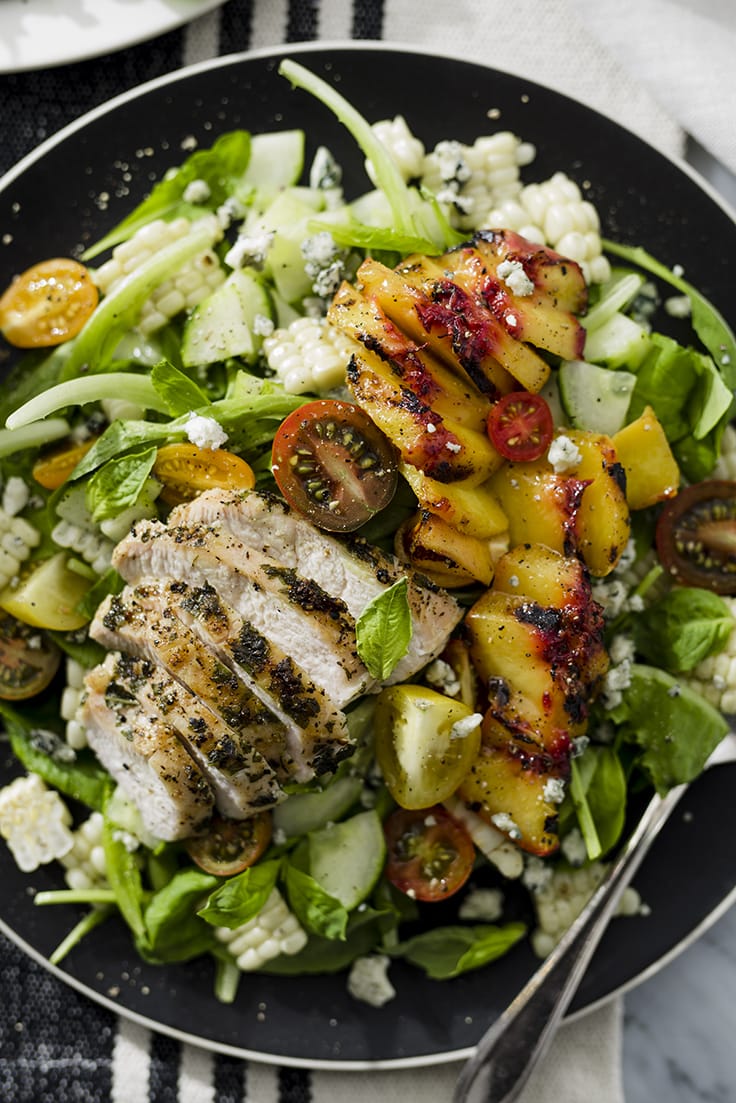 A grilled basil chicken salad with grilled chicken, peaches, and corn over mixed greens with blue cheese crumbles on a black plate.