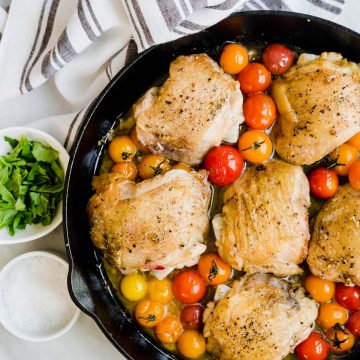 Crispy chicken thighs with burst tomatoes in a cast iron skillet.