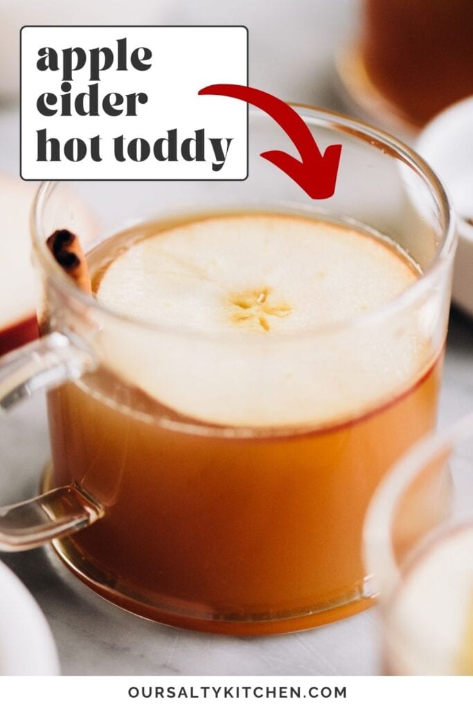 Side view, an apple cider hot toddy in a glass mug on a marble table, garnished with an apple slice and cinnamon stick; text overlay at the top reads "apple cider hot toddy".