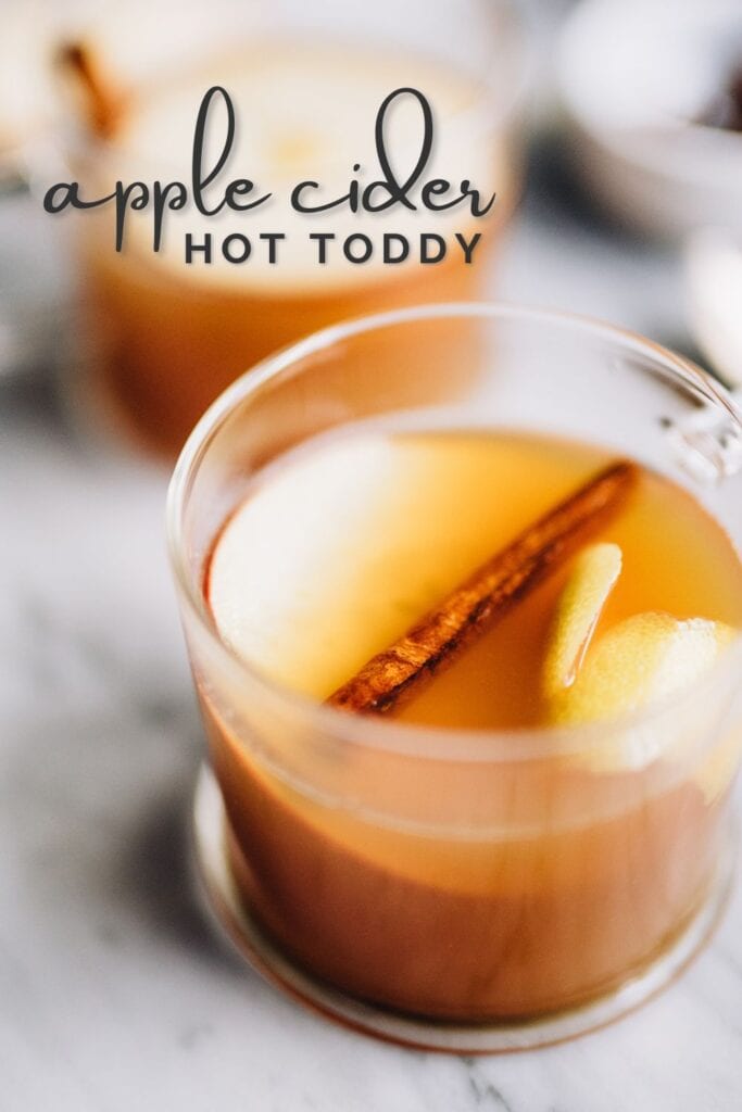 Side view, two apple cider hot toddy cocktails in glass mugs, garnished with apple slices and a cinnamon stick; text overlay at the top reads "apple cider hot toddy".
