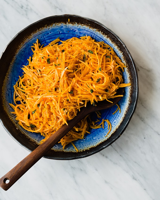A large bowl of vibrant roasted butternut squash noodles tossed with crispy sage butter with a wood serving spoon on a marble table. 