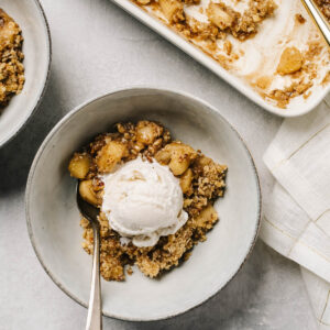 A bowl of gluten free apple crisp topped with vanilla ice cream on a table with a white linen napkin.