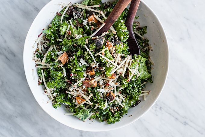 A large white salad bowl filled with the base of kale chicken salad - chopped kale, apples, quinoa, warm potatoes, and a maple cider vinaigrette. 