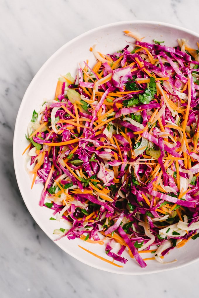 A bowl of apple cider vinegar coleslaw with red cabbage, carrots, fennel and parsley on a marble table. 