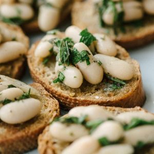 This recipe for rosemary marinated white bean bruschetta is the low-on-prep, high-on-taste appetizer of your cocktail party dreams. This recipe is vegan and vegetarian, incredibly easy, requires no fancy kitchen equipment, and needs just 15 minutes of prep. Who knew beans on toast could be so delicious?!?