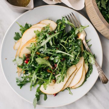 Arugula pear salad with pomegranates and pistachios on a blue plate with a silver fork a small pitcher of champagne mustard vinaigrette.