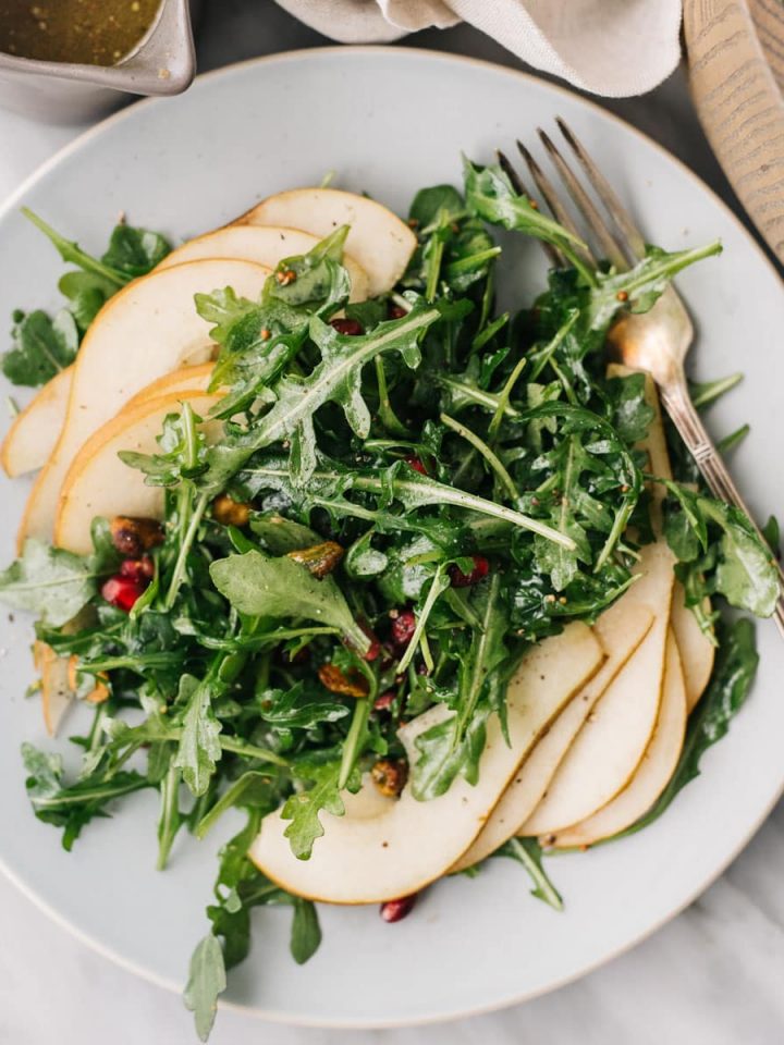Arugula pear salad with pomegranates and pistachios on a blue plate with a silver fork a small pitcher of champagne mustard vinaigrette.