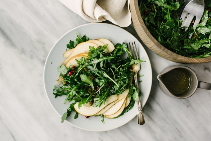 A large wood bowl and an individual serving of arugula pear salad on a marble table.