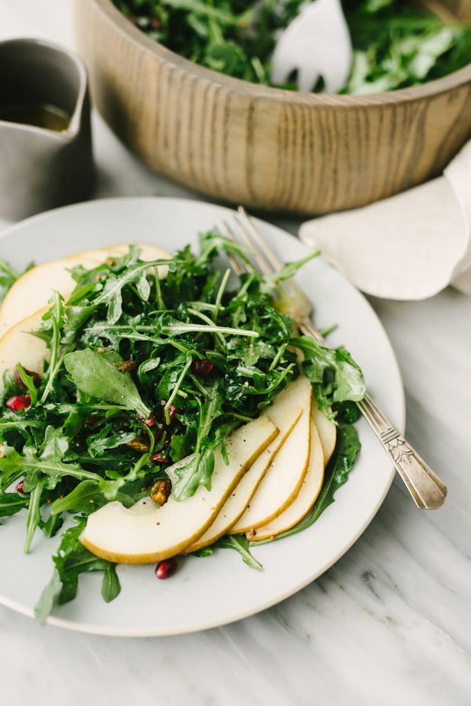 A vegan and whole30 arugula pear salad with pistachio and pomegranate on a blue plate with a small pitcher of vinaigrette on the side.