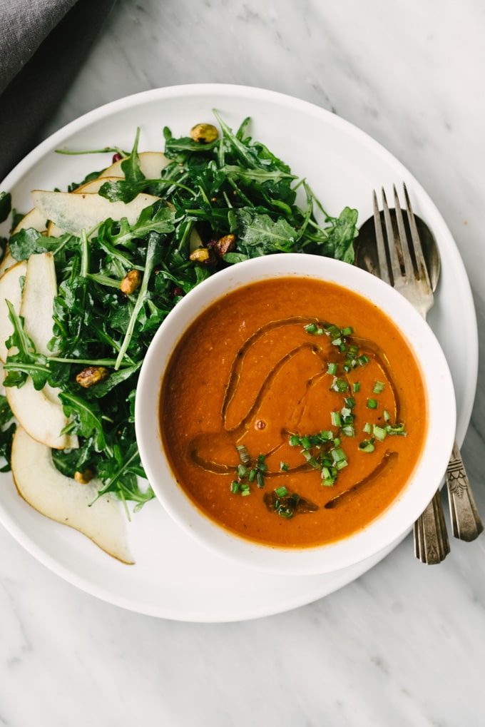 A white plate with a bowl of tomato soup and a side serving of vegan arugula pear salad.