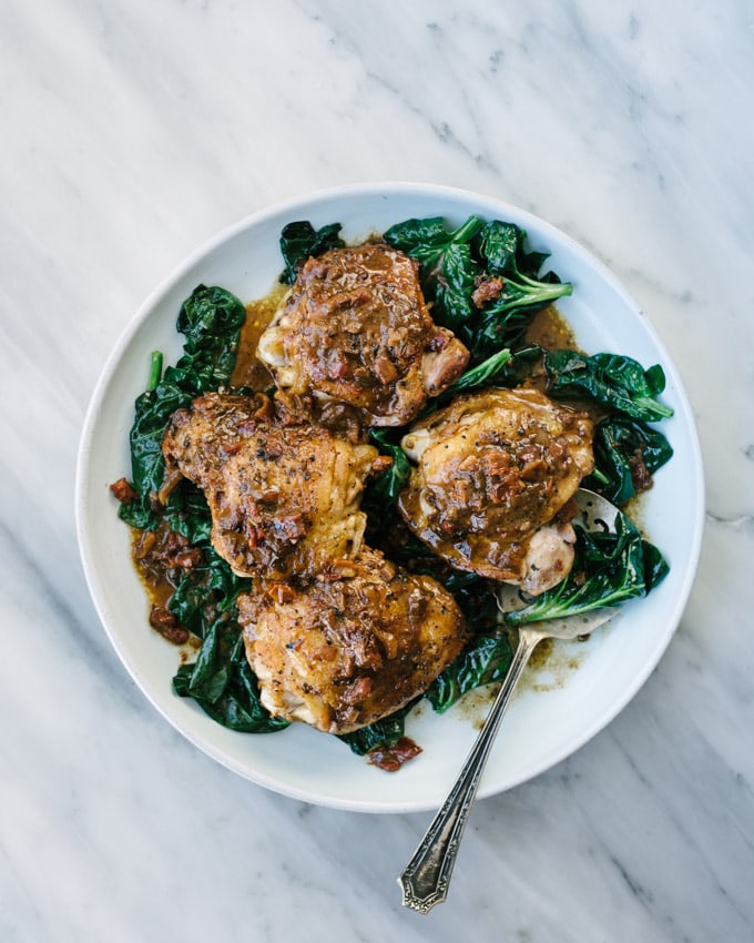 A platter of crispy and cream sun dried tomato tomato chicken thighs over a bed of wilted spinach on a marble table. 
