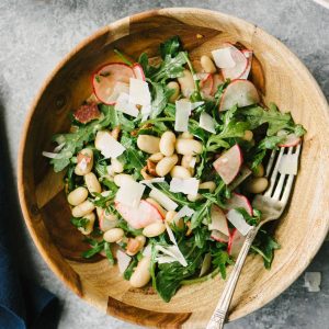 Gluten free and grain free white bean salad with bacon, radishes, fresh herbs, and arugula in a wooden bowl on a cement background.