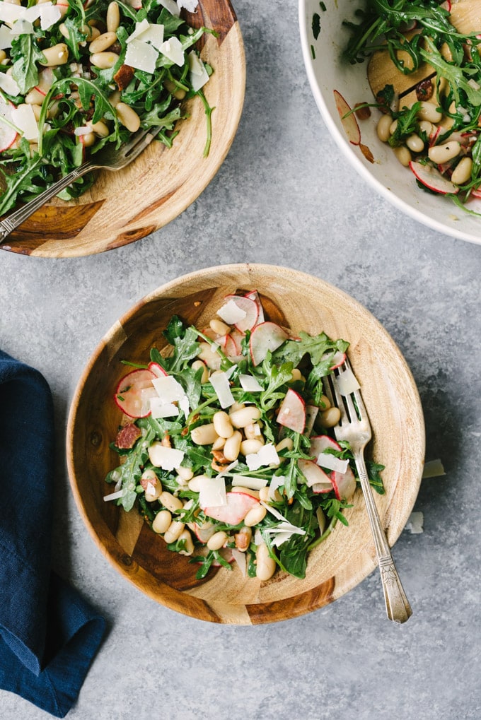 Gluten free and grain free white bean salad with bacon, radishes, fresh herbs, and arugula in a wooden bowl on a cement background. 