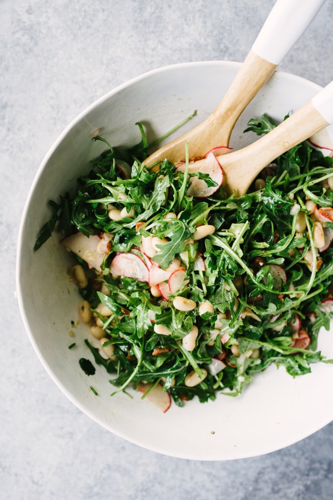 Marinated white bean salad gently tossed with fresh arugula in a large white salad bowl.