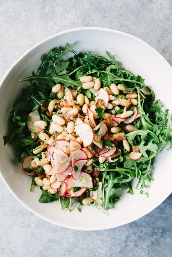 Marinated white bean salad on top of fresh arugula leaves in a large white salad bowl.