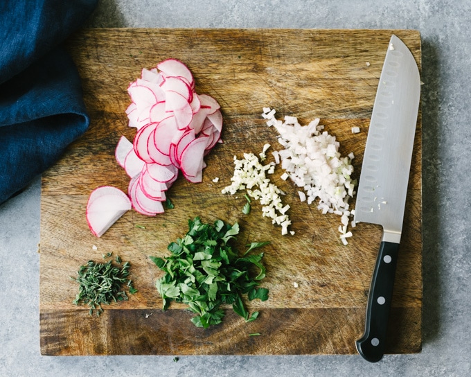 How to make white bean salad. Sliced radishes, chopped herbs, and minced shallot and garlic on a wooden cutting board.