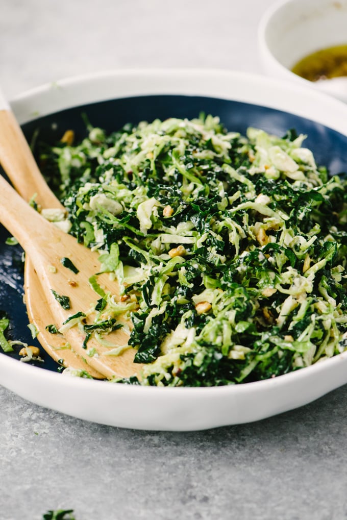 A bowl of kale and brussels sprouts salad with wood serving tongs on a cement table with a small bowl of hazelnut vinaigrette in the background.