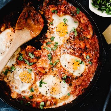 Gluten free white bean eggs in purgatory in a cast iron skillet with herbs and parmesan cheese on the side.