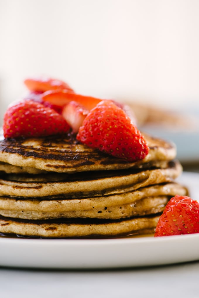 A detail shot of a stack of whole grain pancakes on a white plate topped with macerated strawberries and a drizzle of real maple syrup.