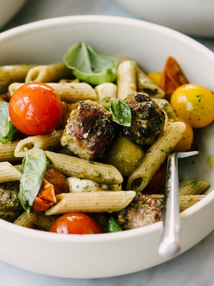 A bowl of caprese pasta tossed with Italian sausage and clean basil pesto on a marble table.
