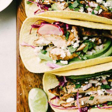 Three grilled chicken tacos with avocado, cilantro, and lime on a cutting board.