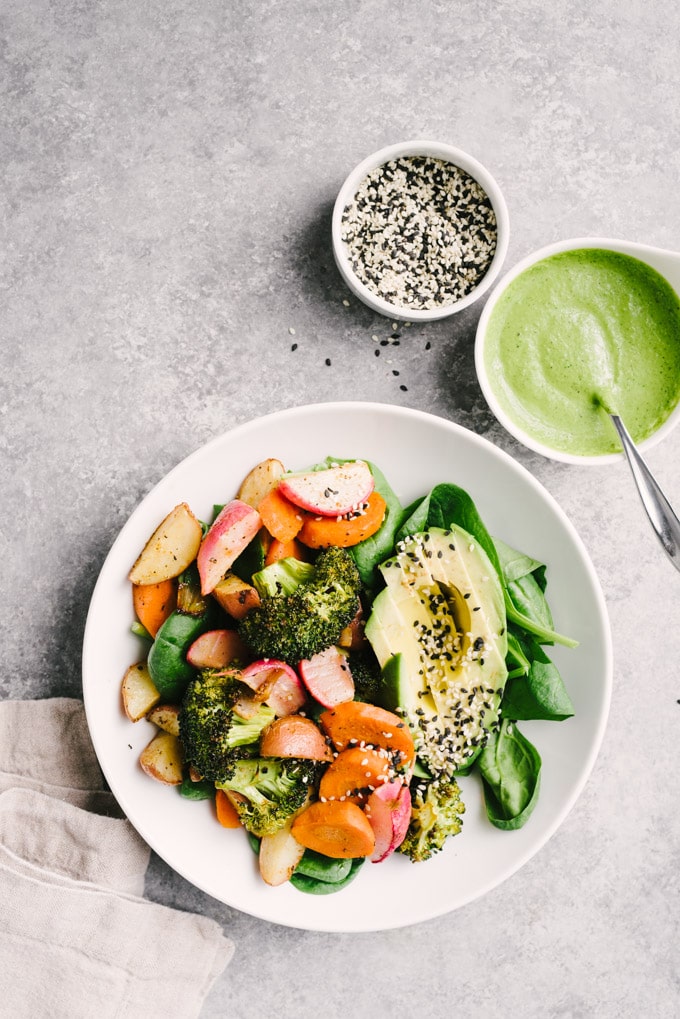 An overhead view of a roasted vegetable salad served with half an avocado, sesame seeds, and a side dish of green tahini sauce on a concrete table.