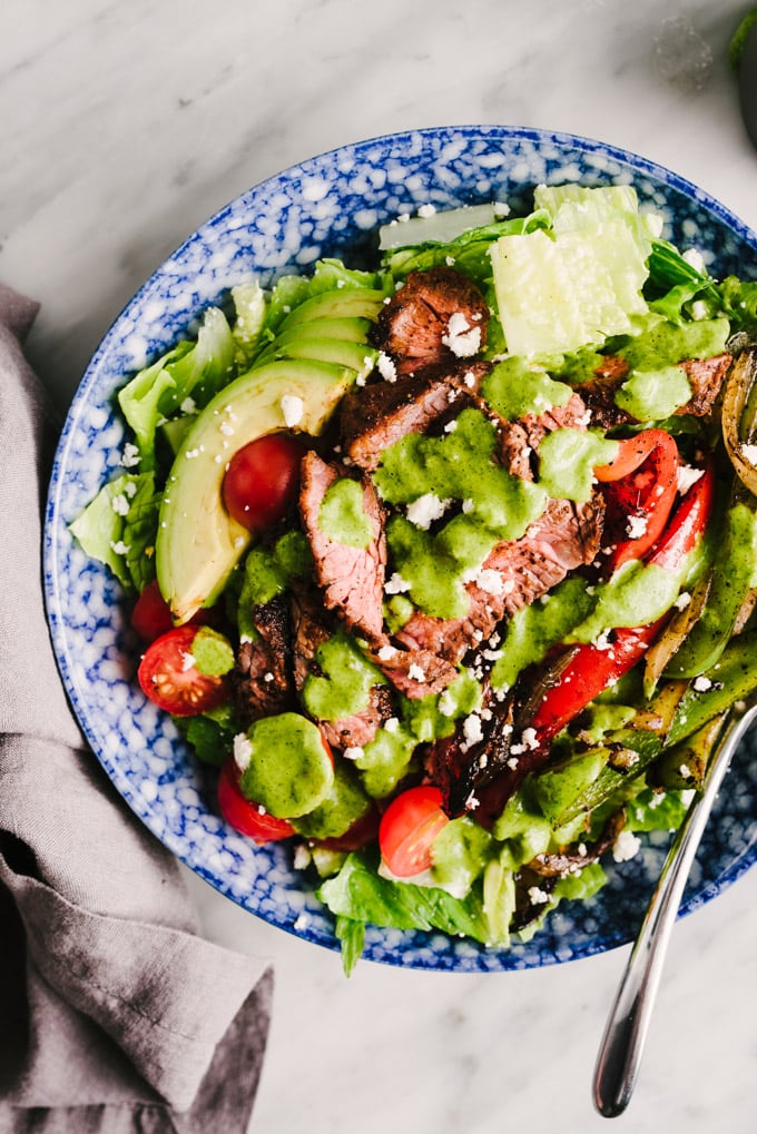 Steak fajita salad drizzled with cilantro lime dressing in a blue and white bowl on a marble table.