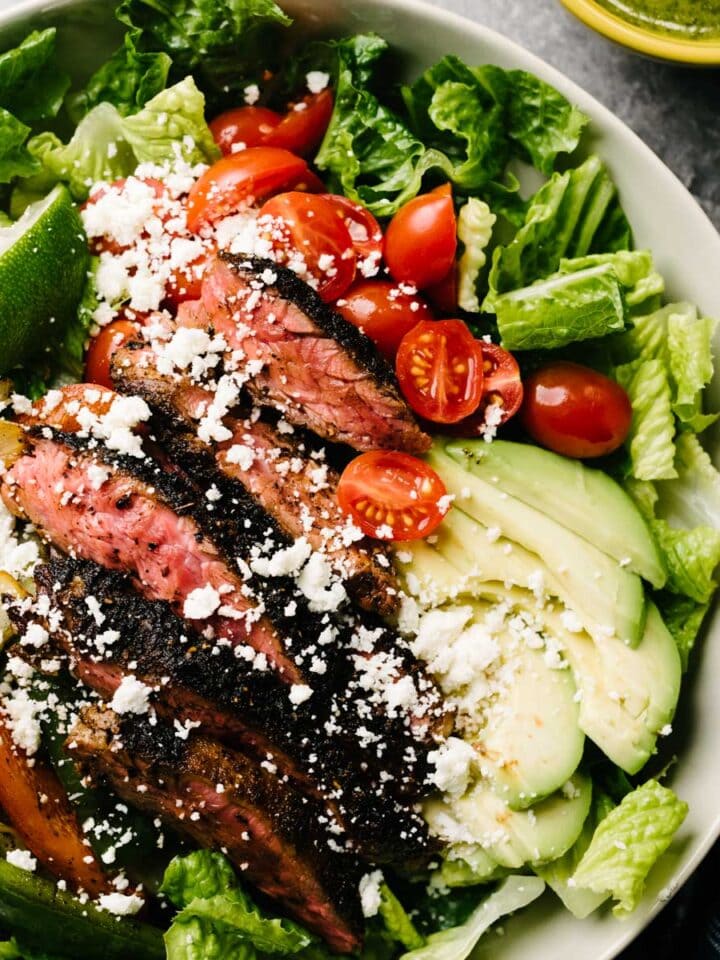 Steak fajita salad in a low tan serving bowl, made with chopped romaine, tomatoes, and avocado topped with seared fajita steak and sautéed peppers and onions.