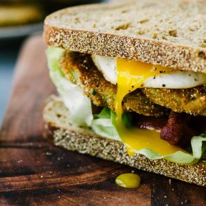 A fried green tomato BLT sandwich on whole grain bread with a fried egg on a wood cutting board.