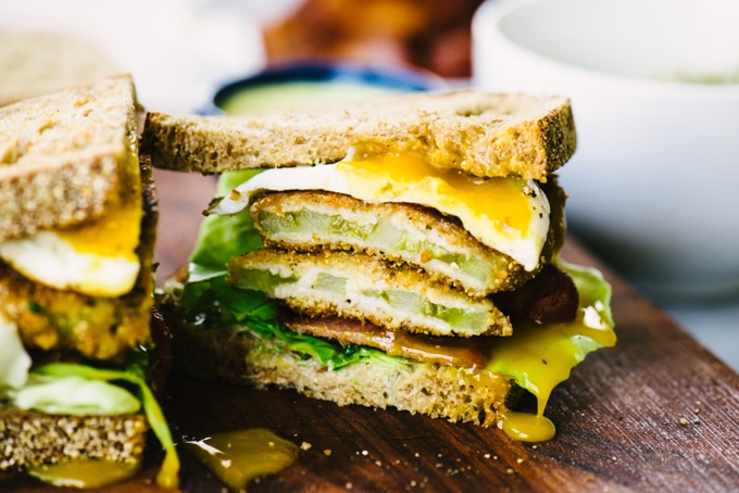 A fried green tomato BLT sandwich sliced in half with a fried egg on a wood cutting board.