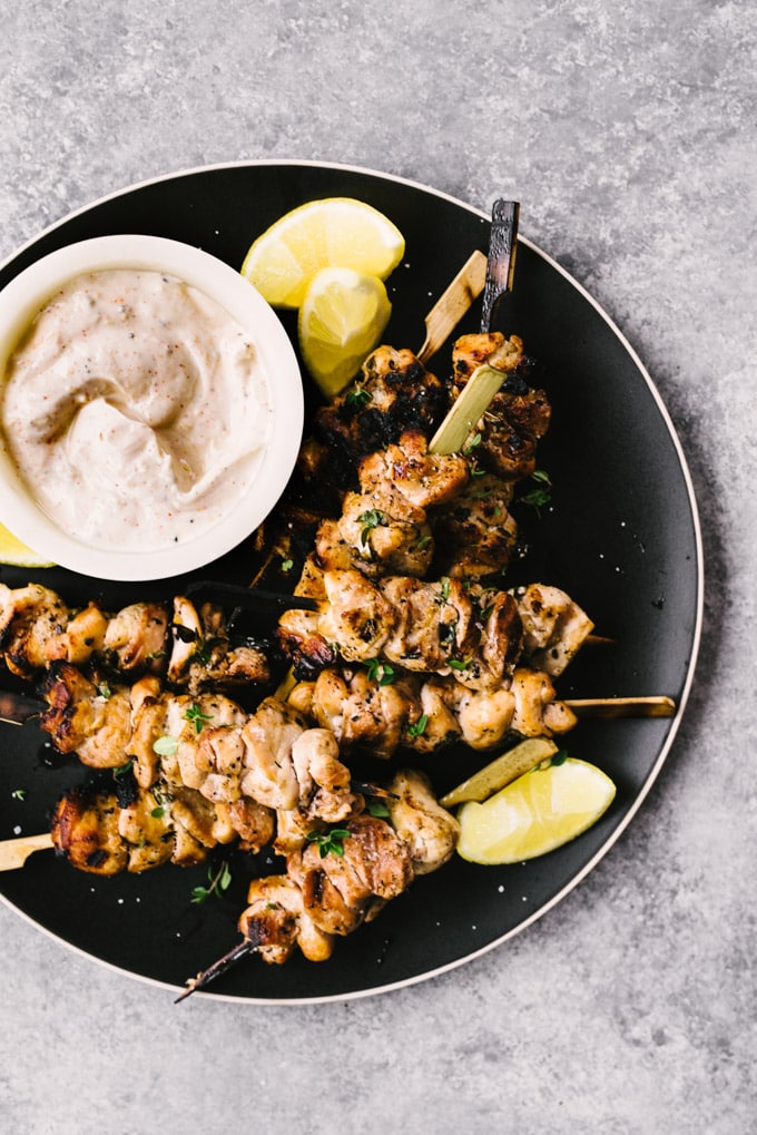 A large black serving dish filled with grilled lemon thyme chicken skewers, lemon wedges, and a small white bowl of lemon yogurt dipping sauce.