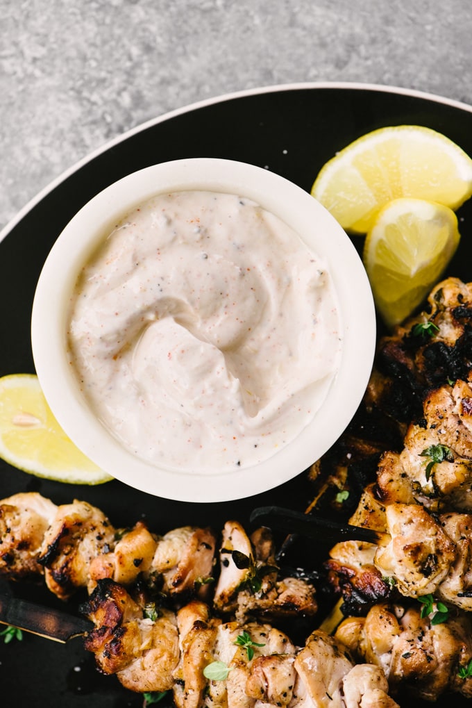 Yogurt dip with lemon and paprika on a black plate surrounded by lemon wedges and lemon thyme chicken skewers.