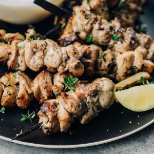 Grilled lemon thyme chicken skewers on a black plate with lemon wedges.