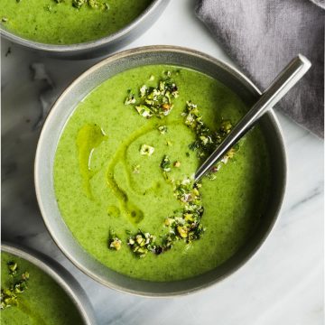 A bowl of green spinach potato soup on a marble table with a grey napkin and a small bowl of pistachio gremolata.