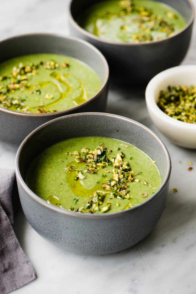 Three small bowls of whole30 and paleo potato spinach soup on a marble table with a small dish of pistachio gremolata on the side.