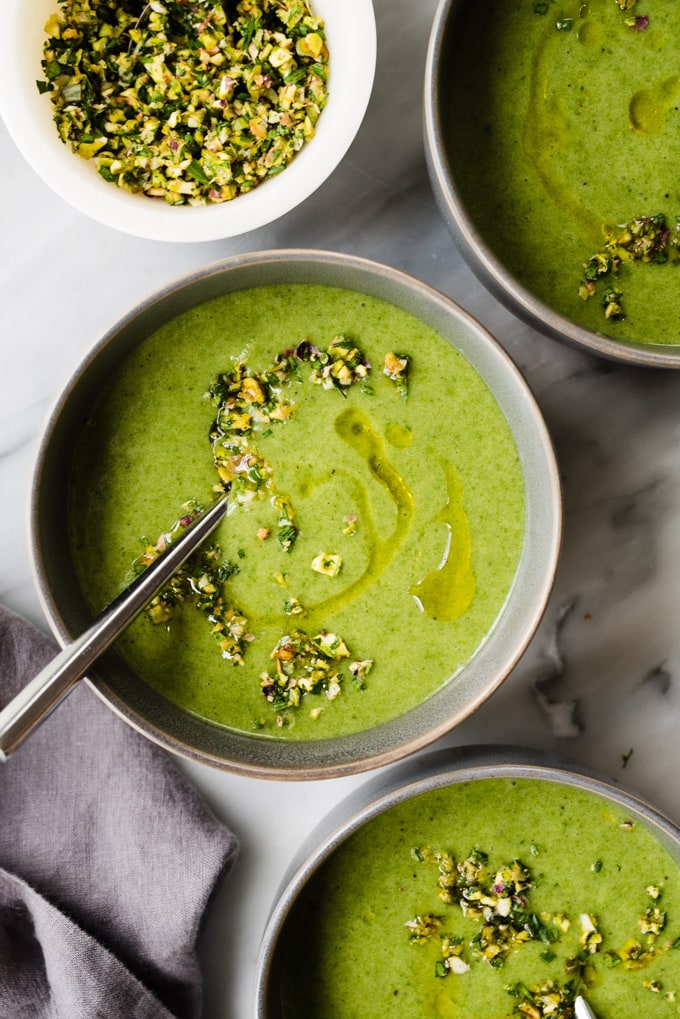 Three bowls of green spinach potato soup on a marble table with a grey napkin and a small bowl of pistachio gremolata.