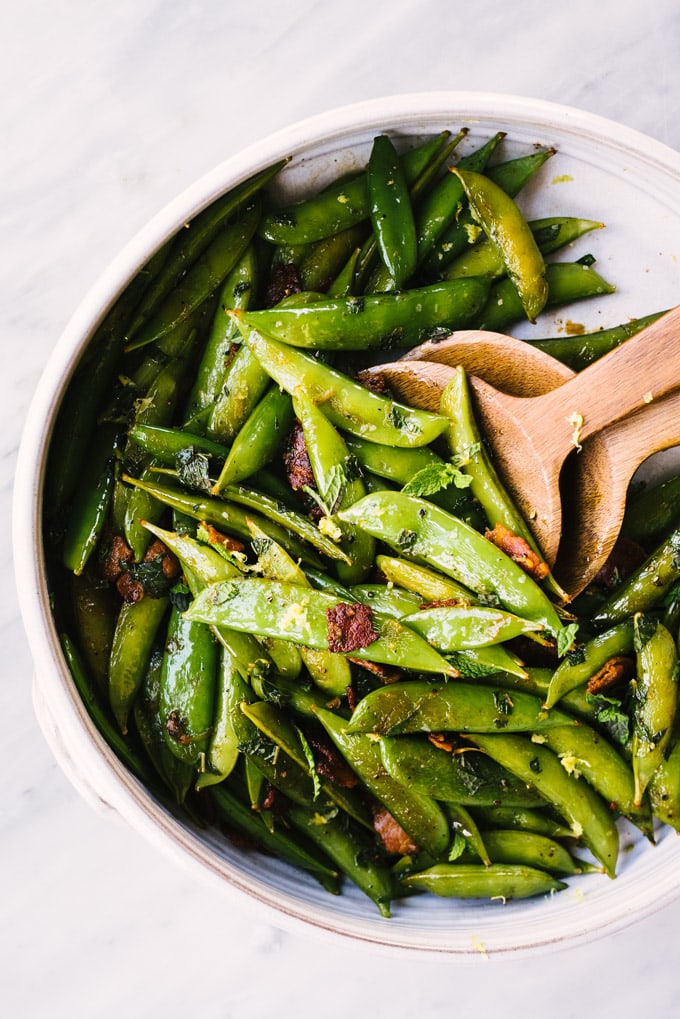A recipe for sugar snap peas cooked with bacon, mint, and lemon in a large serve bowl with wooden serving utensils.