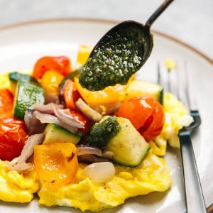 Whole30 pesto sauce being drizzled over scrambled eggs topped with roasted vegetables.
