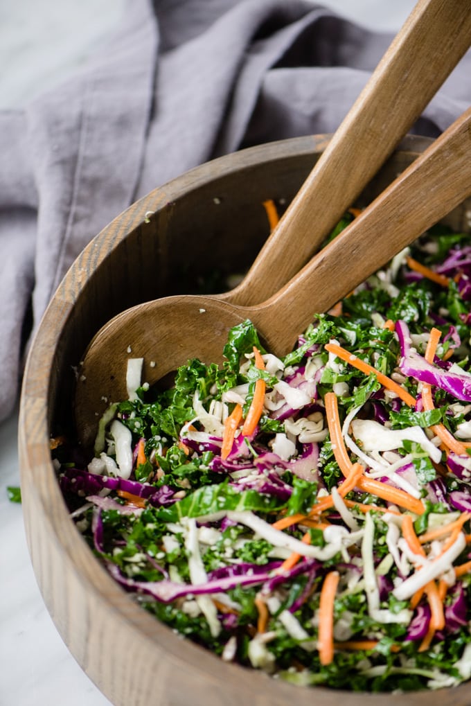 A wood serving bowl with serving spoons filled with colorful, crunchy kale slaw.